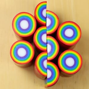 May include: A close-up of eight rainbow-colored polymer clay canes, each with a white center, arranged in a circular pattern on a wooden surface.