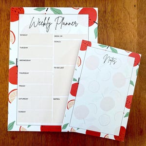 May include: A weekly planner and a notes pad featuring a white background with a pattern of red apples and green leaves. The planner includes sections for Monday through Sunday, goals, and a to-do list. The notes pad has a blank lined area for writing.