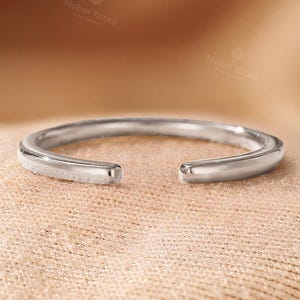 May include: A silver-colored open ring with rounded edges. The ring is set against a neutral, textured background. The ring is a simple, elegant piece of jewelry.
