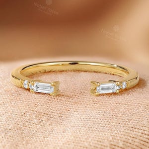 May include: A gold-colored open ring with two baguette-cut diamonds and small round diamonds. The ring is set on a peach-colored surface. The brand name "Nobel Yates" is visible in the background.