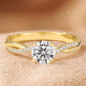 May include: A gold engagement ring with a round, clear center stone. The band features a twisted design with small, sparkling accent stones. The ring is set against a soft, peach-colored background.