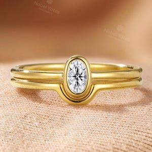Pode incluir: Um conjunto de anéis dourados com um diamante de corte oval. O anel principal apresenta uma cravação solitária, com uma segunda banda por baixo. O fundo é de cor neutra.