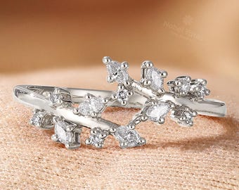 May include: A silver ring with a leaf-like design, adorned with multiple clear, sparkling gemstones. The ring's band is thin, and the gemstones are arranged to resemble leaves and branches. The ring is displayed on a peach-colored fabric.