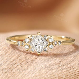 May include: A gold ring featuring a square-cut, clear gemstone at its centre. The ring is embellished with smaller, round gemstones on either side of the main stone. The ring is set against a soft, peach-coloured background. The ring is from Nobel Yates Fine Jewellery.