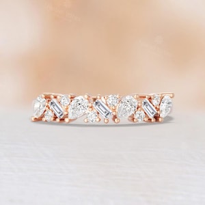 May include: A rose gold ring with alternating pear-shaped and baguette-cut clear gemstones. The gemstones are set closely together, creating a continuous, sparkling band. The ring is displayed on a white surface with a soft, blurred background.