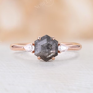 Pode incluir: Um anel de noivado de ouro rosa com um diamante hexagonal cinza e dois diamantes de acentua&ccedil;&atilde;o em forma de p&ecirc;ra.