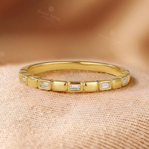May include: A gold band ring featuring rectangular gemstones set around the band. The ring is presented on peach-coloured fabric. The image includes the text "Nobel Yates" on the top left.