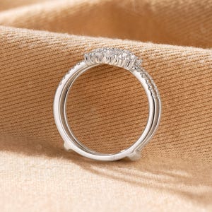 May include: A silver-toned ring guard featuring a curved bridge set with small, clear, round-cut stones. The band consists of two parallel metal shanks that meet at the top, creating an open space designed to accommodate another ring.