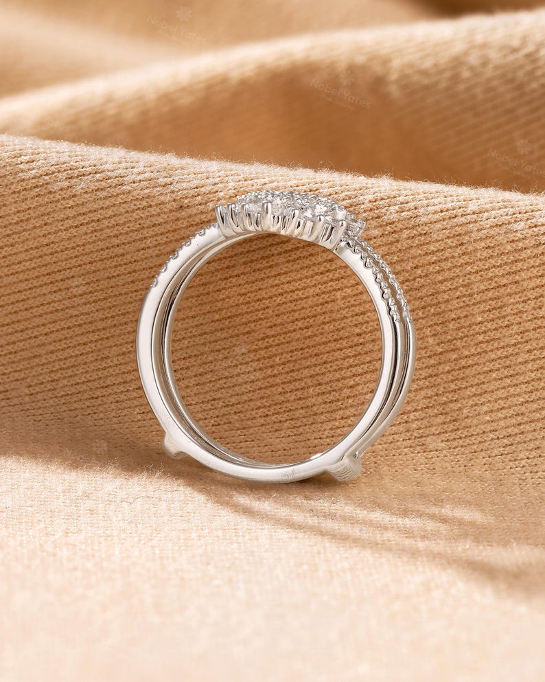 May include: A silver-toned ring guard featuring a curved bridge set with small, clear, round-cut stones. The band consists of two parallel metal shanks that meet at the top, creating an open space designed to accommodate another ring.