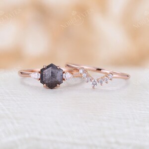 Pode incluir: Um anel de noivado de ouro rosa com um diamante negro hexagonal e dois diamantes de acentua&ccedil;&atilde;o em forma de p&ecirc;ra. A alian&ccedil;a de casamento correspondente apresenta uma fila delicada de diamantes redondos em um design curvo.