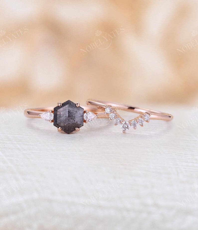 Pode incluir: Um anel de noivado de ouro rosa com um diamante negro hexagonal e dois diamantes de acentua&ccedil;&atilde;o em forma de p&ecirc;ra. A alian&ccedil;a de casamento correspondente apresenta uma fila delicada de diamantes redondos em um design curvo.