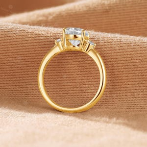 May include: A gold ring featuring a large, round, clear center stone held by four prongs, flanked by two smaller, round, clear side stones. The band is smooth and polished, showcasing a classic design with a prominent setting for the main gemstone.