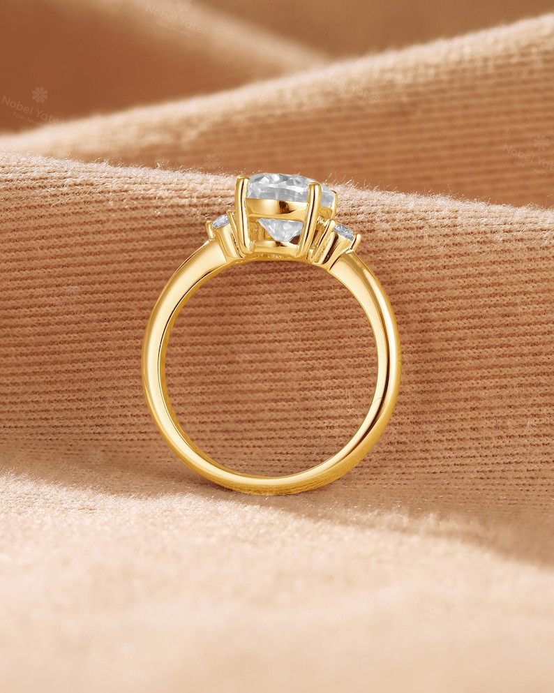 May include: A gold ring featuring a large, round, clear center stone held by four prongs, flanked by two smaller, round, clear side stones. The band is smooth and polished, showcasing a classic design with a prominent setting for the main gemstone.