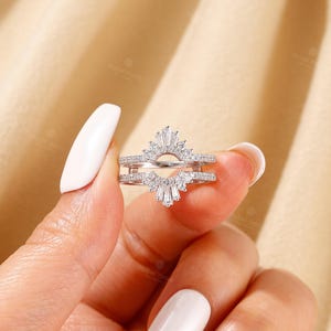 May include: A silver-toned ring guard featuring two bands connected by a central arch. The design is encrusted with small round stones and accented with baguette-cut stones arranged in a fan-like pattern at the top and bottom of the arch.