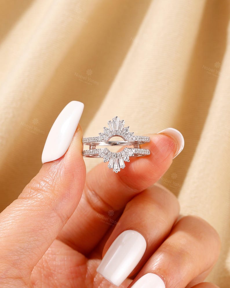 May include: A silver-toned ring guard featuring two bands connected by a central arch. The design is encrusted with small round stones and accented with baguette-cut stones arranged in a fan-like pattern at the top and bottom of the arch.