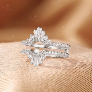May include: A silver-toned ring guard featuring two arched bands connected by a vertical bar. The design includes a central fan-shaped arrangement of baguette-cut stones flanked by round-cut stones, with additional round stones set along the bands.