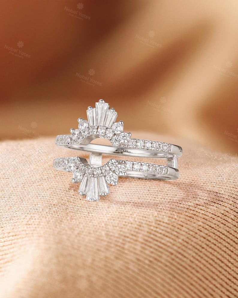 May include: A silver-toned ring guard featuring two arched bands connected by a vertical bar. The design includes a central fan-shaped arrangement of baguette-cut stones flanked by round-cut stones, with additional round stones set along the bands.
