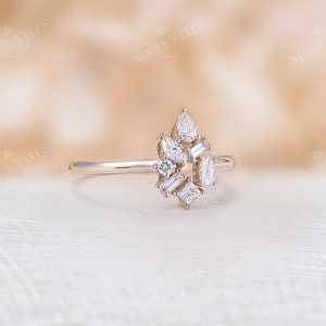 May include: A rose gold ring with a cluster of diamonds in different shapes. The diamonds include pear, baguette, and square cuts, arranged in a floral or starburst pattern. The ring has a slender band and a polished finish.