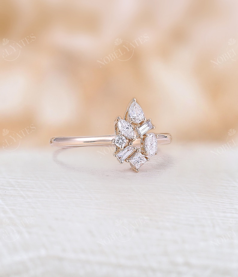 May include: A rose gold ring with a cluster of diamonds in different shapes. The diamonds include pear, baguette, and square cuts, arranged in a floral or starburst pattern. The ring has a slender band and a polished finish.