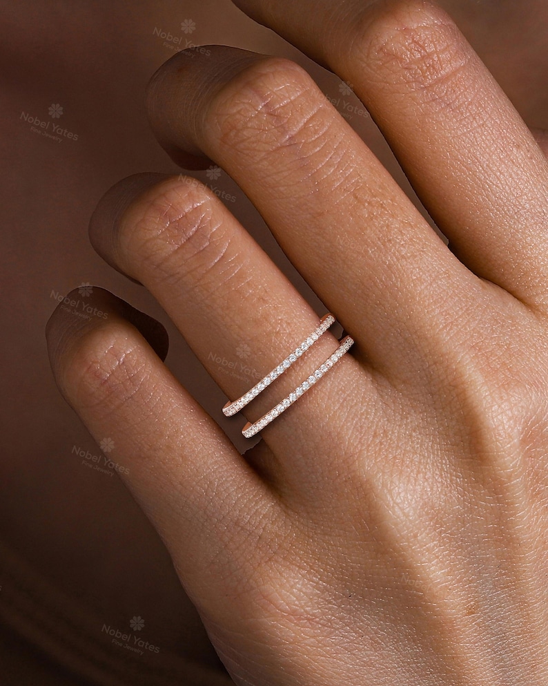 May include: A rose gold double band ring featuring two parallel rows of small, clear, round-cut pav&eacute;-set stones. The ring has an open design, with the two bands connected at the back, creating a layered appearance across the finger.