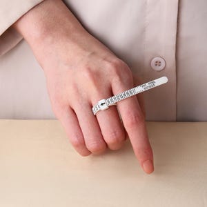May include: A white plastic ring sizer tool featuring a numbered scale from 1 to 17, with a black arrow indicator and the text PAT. PEND. 9101693 printed on the band. The adjustable strap is looped around a finger to determine ring size.