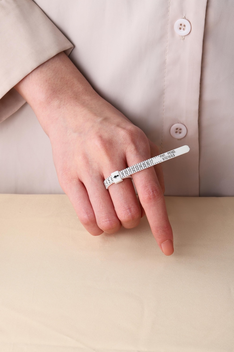May include: A white plastic ring sizer tool featuring a numbered scale from 1 to 17, with a black arrow indicator and the text PAT. PEND. 9101693 printed on the band. The adjustable strap is looped around a finger to determine ring size.