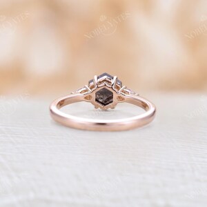 Pode incluir: Um anel de noivado de ouro rosa com uma pedra central de diamante hexagonal cinza e dois diamantes menores de cada lado.
