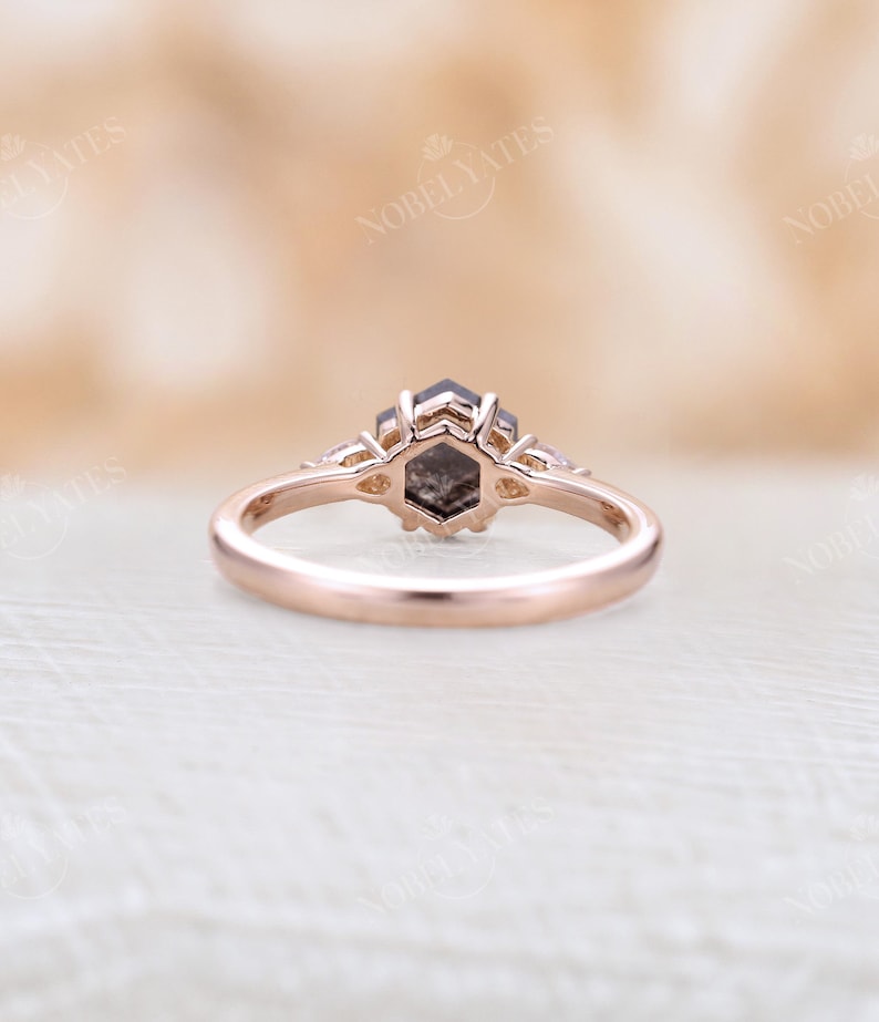 Pode incluir: Um anel de noivado de ouro rosa com uma pedra central de diamante hexagonal cinza e dois diamantes menores de cada lado.