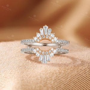 May include: A double-band ring guard featuring a sunburst design with clear baguette and round-cut stones set in rose gold. The two bands are connected by a curved bridge, with pav&eacute;-set stones along the shanks and a central fan-like arrangement.