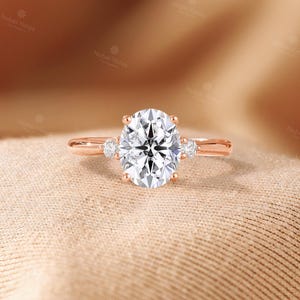 May include: A rose gold ring featuring a large oval-cut center stone held by four prongs, flanked by a single smaller round-cut stone on each side. The band is smooth and polished, showcasing a classic three-stone design with clear, faceted gems.