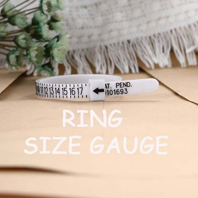 May include: A white plastic ring size gauge featuring black numerical markings from 10 to 17 and a sliding buckle with an arrow indicator. The text AT. PEND. 101693 is printed on the strap. The tool is used for measuring finger circumference.
