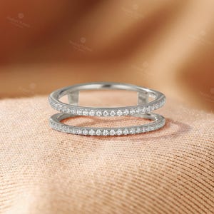 May include: A silver-toned double band ring featuring two parallel bands connected by vertical bars. Both bands are encrusted with a continuous row of small, clear, round-cut stones that catch the light, creating a sparkling, uniform appearance.