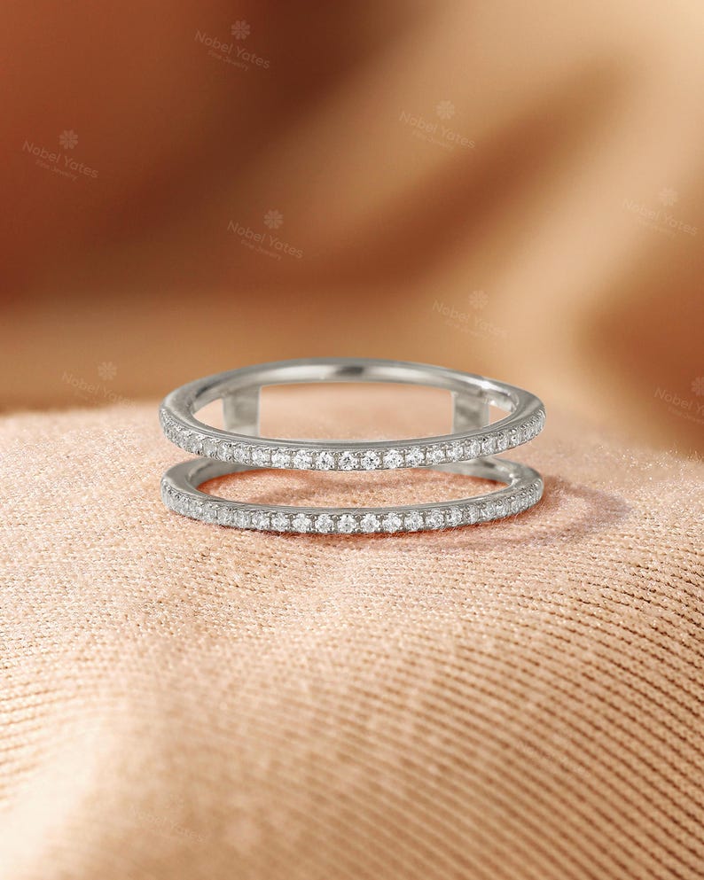 May include: A silver-toned double band ring featuring two parallel bands connected by vertical bars. Both bands are encrusted with a continuous row of small, clear, round-cut stones that catch the light, creating a sparkling, uniform appearance.