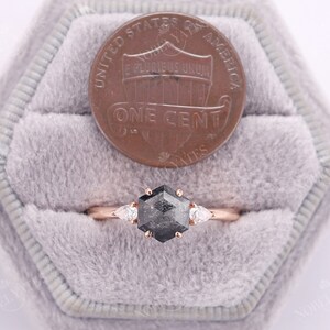 Pode incluir: Um anel de noivado de ouro rosa com um diamante preto hexagonal e dois diamantes em forma de p&ecirc;ra de cada lado. O anel est&aacute; em uma pequena caixa de veludo com um centavo americano para refer&ecirc;ncia de tamanho.