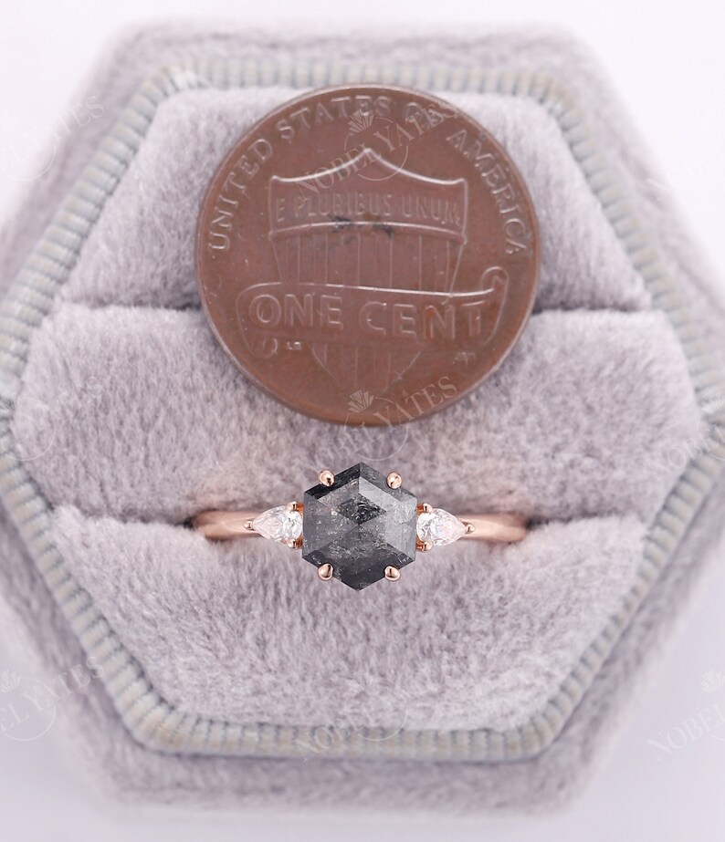 Pode incluir: Um anel de noivado de ouro rosa com um diamante preto hexagonal e dois diamantes em forma de p&ecirc;ra de cada lado. O anel est&aacute; em uma pequena caixa de veludo com um centavo americano para refer&ecirc;ncia de tamanho.