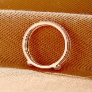 May include: A rose gold ring guard featuring a curved, open-ended design with a polished metal finish. The outer band is adorned with a row of small, clear pav&eacute;-set stones that extend along the top surface, creating a delicate and refined appearance.