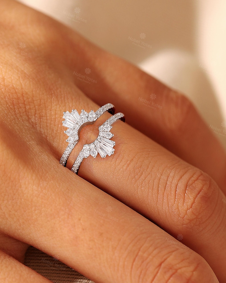 May include: A silver-toned ring guard featuring a curved design adorned with multiple small round-cut stones and two prominent baguette-cut stones at the ends. The band is encrusted with tiny pav&eacute;-set stones along the sides, creating a sparkling effect.