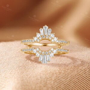May include: A gold ring guard featuring two bands connected by a central arch. The design includes baguette-cut and round-cut clear stones set along the top and bottom edges, creating a symmetrical, sunburst-inspired pattern with a polished finish.