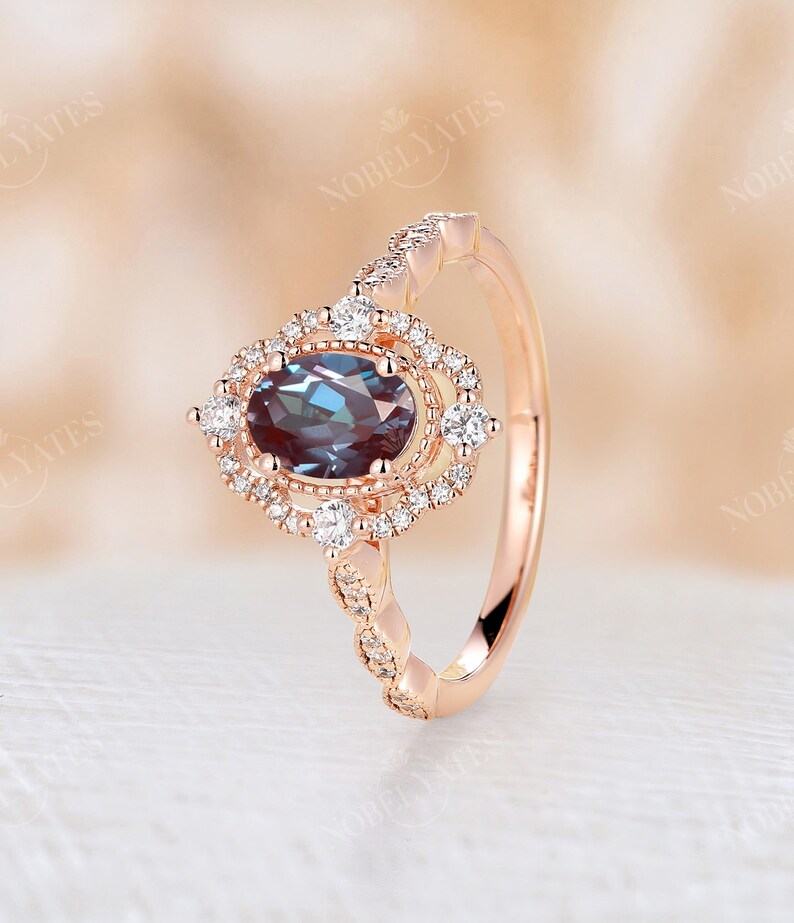 May include: A rose gold engagement ring with a large oval-shaped blue gemstone surrounded by a halo of smaller diamonds. The band is adorned with a row of smaller diamonds.