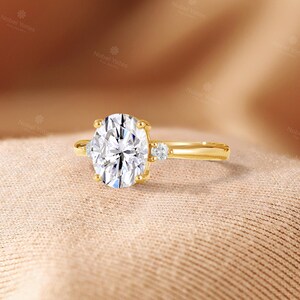 May include: A gold ring featuring a large oval-cut center stone held by four prongs, flanked by a single small round-cut stone on each side of the band. The metal band is smooth and polished, showcasing a classic three-stone design with clear gems.