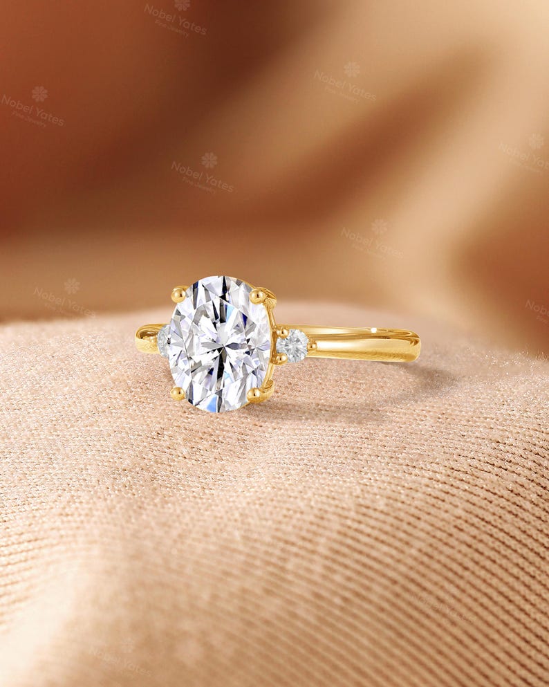 May include: A gold ring featuring a large oval-cut center stone held by four prongs, flanked by a single small round-cut stone on each side of the band. The metal band is smooth and polished, showcasing a classic three-stone design with clear gems.