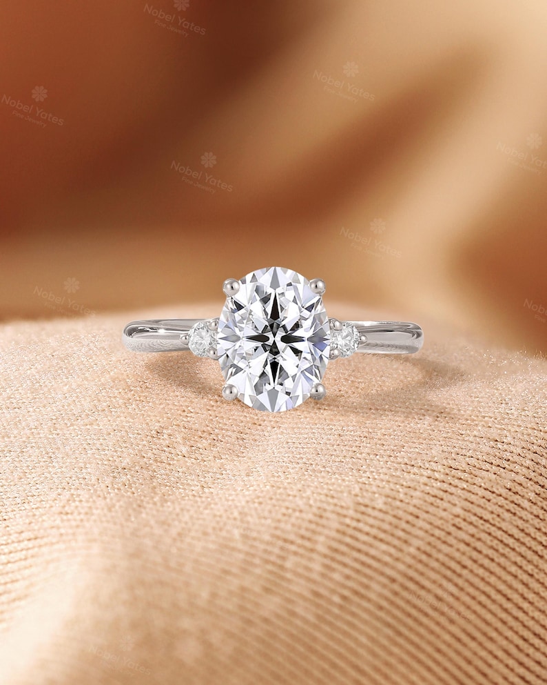May include: A silver-toned ring featuring a large, oval-cut center stone held by four prongs, flanked by two smaller round-cut stones on each side of the smooth, polished band. The jewelry piece displays a clear, faceted design throughout.