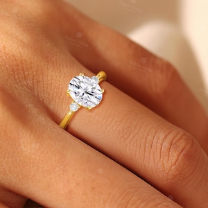 May include: A gold ring featuring a large oval-cut center stone flanked by two smaller round-cut stones. The band is polished yellow gold, and the setting holds the three clear, faceted stones securely in place with metal prongs on a finger.