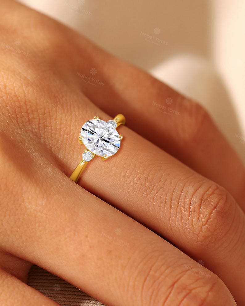 May include: A gold ring featuring a large oval-cut center stone flanked by two smaller round-cut stones. The band is polished yellow gold, and the setting holds the three clear, faceted stones securely in place with metal prongs on a finger.