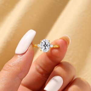 May include: A gold ring featuring a large oval-cut center stone held by four prongs, flanked by two smaller round-cut stones on either side of the band. The metal band is smooth and polished, showcasing a classic three-stone design with clear gems.