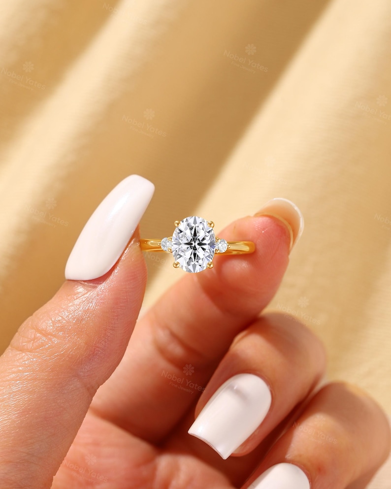 May include: A gold ring featuring a large oval-cut center stone held by four prongs, flanked by two smaller round-cut stones on either side of the band. The metal band is smooth and polished, showcasing a classic three-stone design with clear gems.