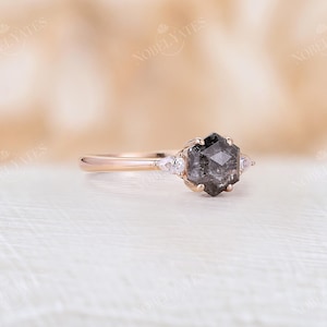 Pode incluir: Um anel de noivado de ouro rosa com uma pedra central de diamante negro hexagonal e dois diamantes menores em forma de p&ecirc;ra de cada lado.