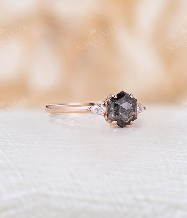 Pode incluir: Um anel de noivado de ouro rosa com uma pedra central de diamante negro hexagonal e dois diamantes menores em forma de p&ecirc;ra de cada lado.