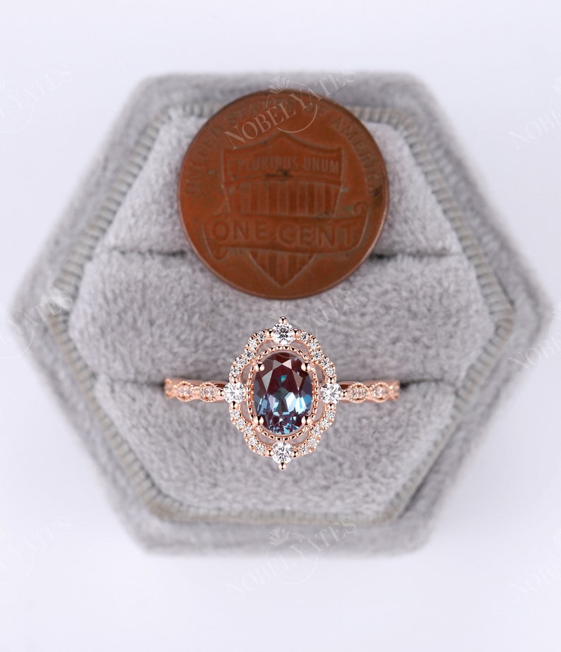 May include: A rose gold ring with a large oval alexandrite gemstone surrounded by a double halo of smaller diamonds. The ring is set in a delicate filigree design and is displayed in a gray velvet ring box.