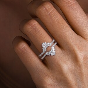 May include: A silver-toned ring guard featuring a curved design with baguette-cut and round-cut clear stones. The band is encrusted with small pav&eacute;-set stones, creating a layered appearance that frames a central space for another ring.